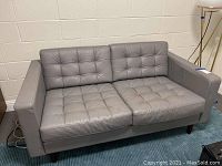 Full view of grey 2-seat sofa with tufted cushions and tapered wooden legs, shown against a wall on a carpeted floor.