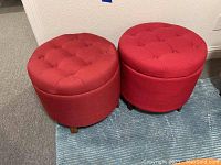 Two round red fabric storage ottomans with button tufted hinged lids, sitting on a blue striped carpet.