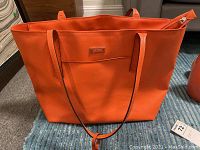 Orange Tumi tote bag photographed standing upright on floor, showing overall front view with straps and logo plaque visible.