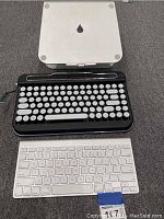 Top view of two keyboards and keyboard stand placed on gray carpet. The retro-style keyboard is black with round white keys and a crank handle. The slim white keyboard is below it. The white keyboard stand with water-drop logo is placed at the back.