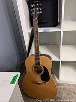 Full view of Mitchell acoustic guitar standing upright showing natural wood finish and black pickguard.