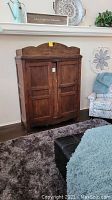 Full front view of the wooden armoire with its two paneled doors and decorative curved top, placed in a living room setting with rug and furniture.