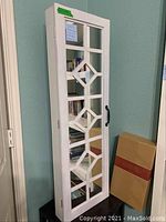 Front upright view of a white wooden jewelry storage cabinet with a mirrored door featuring geometric diamond shapes and a black handle.