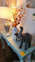 Photo showing a light blue wooden table holding a glass lamp with beige ceramic base and fabric shade, white stoneware cream jug with printed text, decorative artificial orange twigs inside jug, two black elephant figurines, and a small deer figurine.