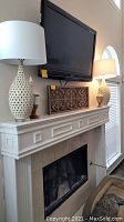 Full view of fireplace mantle with two off-white lattice ceramic lamps with white shades, carved wood wall decor panel below mounted TV, and one wooden urn.