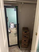 Full view of the black framed wall mirror standing beside the wicker drawer unit.