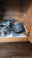 Photo of various pots and pans stacked inside a wood cabinet, including stainless steel and grey non-stick pots with lids and pot holder mat.