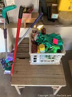 Two wooden benches with a plastic container on top holding multiple cleaning supplies including gloves, spray bottles, and cleaning liquids. Mops with red and blue handles lean beside the benches.