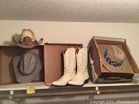 Photo showing three cowboy hats and one pair of white cowboy boots in boxes on a shelf.