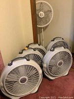 Seven white fans: six floor fans with carry handles and one standing pedestal fan in a corner.