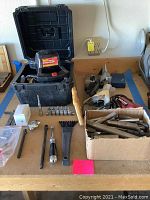 Wide view of garage workbench with assorted tools including speed starter, wrenches, machine parts, and hand saw arranged on top.