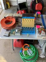 Photo of garage and yard items on white table including orange extension cord, green water hose, blue case with yellow foam, red gas can, action start switch, and other small items.