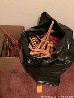 Photo showing a mixed lot of wooden and plastic clothes hangers packed in a black trash bag and a cardboard box, positioned against a wall on a carpeted floor.