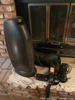 Brown and black Heater brand tower-style heater with video camcorder, lens, and black carrying bag on lace cloth in front of fireplace.