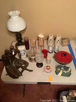 Overview of table displaying white glass oil lamp, metal couple figurine, various candles and holders, decorative door stopper, and red cast iron pot
