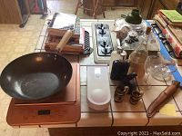 Photo showing Copper Chef wok with wooden handle, digital kitchen scale, stacked cookbooks, white two-burner stovetop, several glass bottles, spice shakers, and other kitchen items on a countertop.