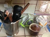 Photo showing metal utensil holder filled with various kitchen utensils, Kitchen Wizard juicer parts, and a brown ceramic lidded container on a kitchen counter near a window.
