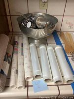 Pewter punch bowl and cups stacked inside, placed behind rolls of cabinet liners and food saver bags.