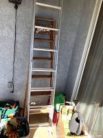 Metal and wooden ladders stacked against a wall, garden gloves and watering cans below them.