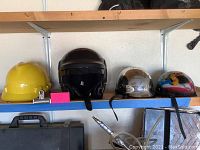Four helmets displayed on a blue shelf including a yellow hard hat, black full-face helmet, dark half helmet, and custom painted helmets.