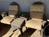 Photo showing two brown plastic patio chairs with palm tree patterned cushions, a small matching side table, and a woven design ottoman arranged outside against a wall.