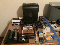 Photo of various office supplies on a table including shredder, calculators, staplers, binding machine, tape dispenser, and staples.