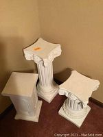 Three white decorative columns arranged in a corner, two with Ionic capitals and one rectangular topped.