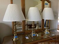 Three brass table lamps with white pleated shades displayed on a wooden surface in front of a mirror.