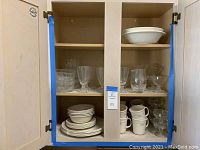 Open kitchen cabinet showing multiple Corelle plates, bowls, mugs, and clear glassware including goblets and short glasses.