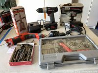 Photo showing the full lot on a white table including Craftsman cordless drill with battery and charger, Black & Decker drill guide box, Black & Decker Handi Saw, Snake Driver Power Pan case, assorted drill bits tray, and boxed Sears electric drill.