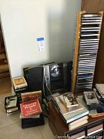 Photo showing Fisher stereo cassette/CD radio with speakers on floor near wooden tower holding multiple CDs, and drawers with cassette tapes and books nearby.