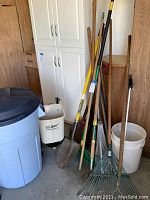 Wide view of gardening tools including shovels, rakes, hoe, axe, broom, trash can, fertilizer spreader, and bucket arranged on garage floor.