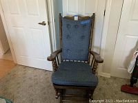 Front view of wooden glider rocking chair with blue cushioned seat and backrest in a room with carpet flooring and white walls.