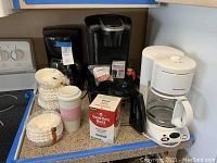 Shows black Keurig coffee maker, black drip coffee maker, white Proctor Silex drip coffee maker with glass carafe, reusable coffee cup, paper coffee filters, box of Seattle's Best coffee pods and Keurig accessories