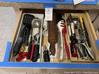 Overhead view of kitchen drawer containing mixed kitchen tools and utensils including wooden rolling pin, spatulas, ladles, peelers, tongs, and small handheld tools.