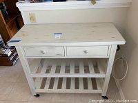 Front view of white distressed serving cart with two drawers, two slatted shelves, and locking casters.