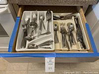 Top view of a kitchen drawer filled with stainless steel forks, spoons, serving spoons, and other utensils organized in white plastic trays.