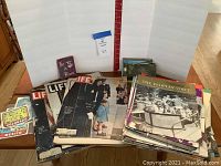 Photo of multiple vintage magazines including Life magazines and The People's Voice magazines stacked and spread out on a table, alongside postcards and decals.