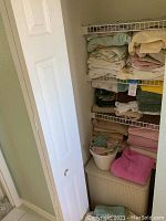 Wide shot of stacking bath towels and bed linens on shelving with laundry basket below.