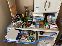 Wide view of office supplies arranged on desk surface and under the desk including mugs with pens, staplers, scissors, calculator, mouse, paper clips, and stationery items.