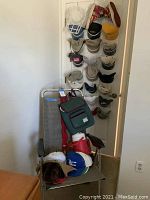 Gray folding outdoor chair with stacked baseball caps and red bag on seat, with baseball cap hanging organizer with 18 hats on door in background.