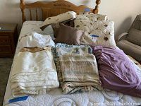Queen size bed covered with folded bedding items including cream and patterned linens, quilt pillow shams, and decorative pillows in brown and purple.