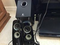 Logitech 3-piece speaker system with subwoofer and satellite speakers shown with Samsung Blu-ray DVD player and Sony TV corner visible.