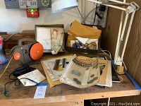 Photo showing Glastar foiling machine, stained glass pattern books, white adjustable work light and soldering tools spread on wood surface.