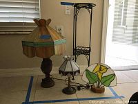 Three stained glass lamps and a metal plant stand with glass pressed flower top are displayed together. Shows all lamps and table in one view.
