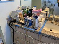 Bathroom countertop with various items including heating pad, blood pressure monitor, wig in stand, curling irons, and personal care accessories