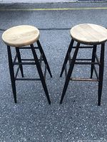Two tall solid wood bar stools with round natural wood seats and black painted legs, shown standing on asphalt.