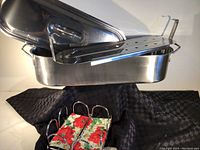 Three-piece stainless steel oval roaster set including pan, lid, and insert, displayed with black checkered tablecloth and napkin holder with floral napkin.