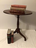 Full view of the round wooden pedestal table with three curved legs and five vintage books placed on and beside it.