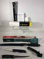 Bushnell riflescope in box with paper instructions, Buck hunting knife in sheath with box, Normark and fillet knives, Scotty sharpener visible